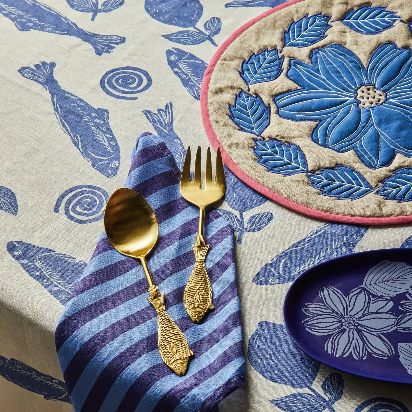 Bonnie and Neil Margot Blue Placemats, napkin with bold blue and purple stripes, on which gold-toned utensils with fish-shaped handles are placed.