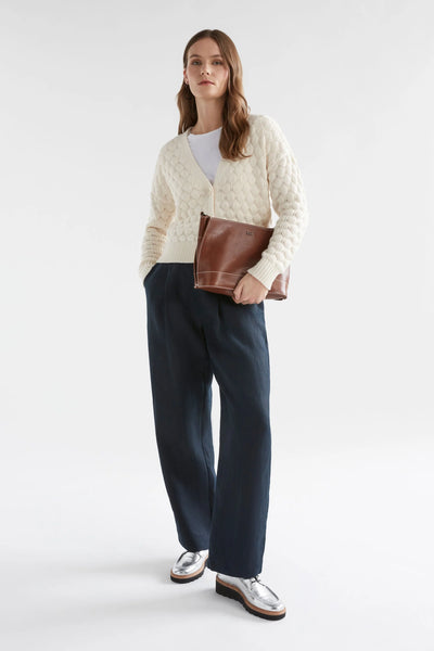 Woman standing wearing black pants, white top with emmer cardigan in oatmeal colour, holding brown clutch bag