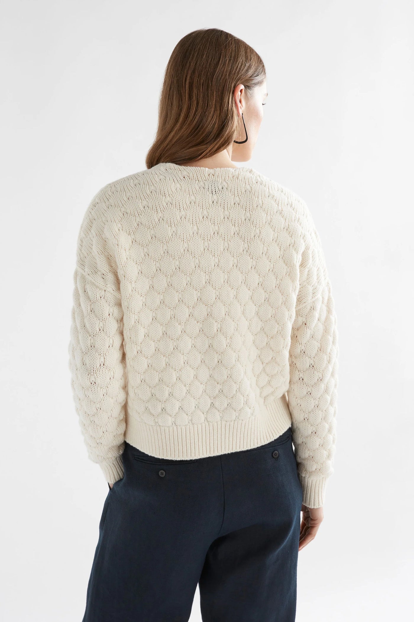 Woman facing the back wearing black pants, white top with emmer cardigan in oatmeal colour