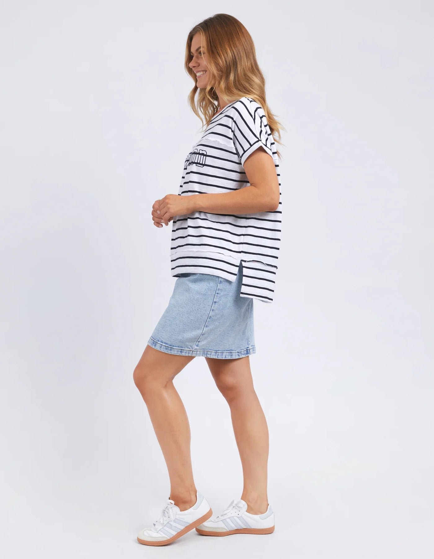 side view of a standing woman wearing foxwood throw on tee white navy stripe, denim skirt and samba shoes