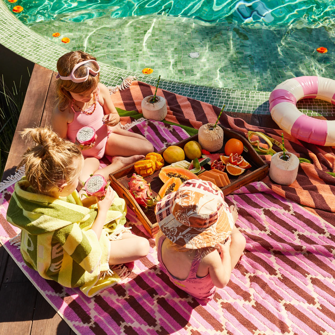 three kids eating fruits beside the pool, one of them is wearing sage and clare jarita hooded towel in palm colour